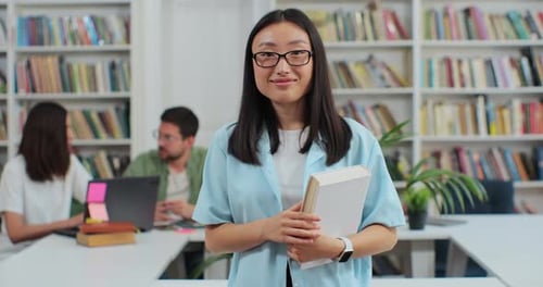 Portrait of Smart Beautiful Young Asian Girl in Glasses Holding Book in Library Bookshelf Background