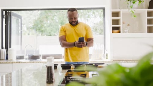 African American man in yellow tee at kitchen island checking phone, tapping 3 meals for timing