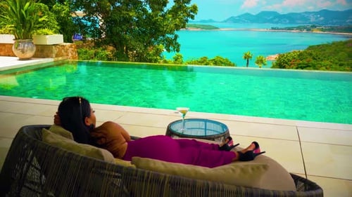 a woman with a sexy evening dress on a trip sitting on a sofa by the pool drinking and looking at th