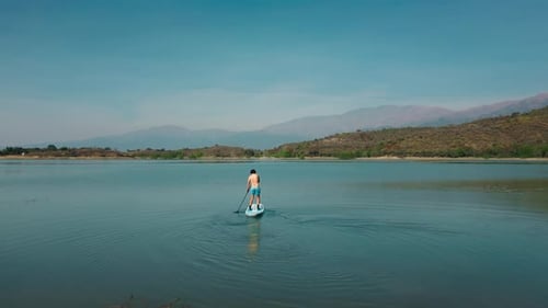 Aerial drone flying over a man doing stand up paddle in a blue water lake with mountains