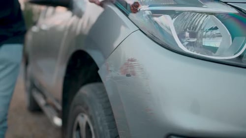 Man touches the damaged front of a car's paint after the car crashes into a parked car on a nearby