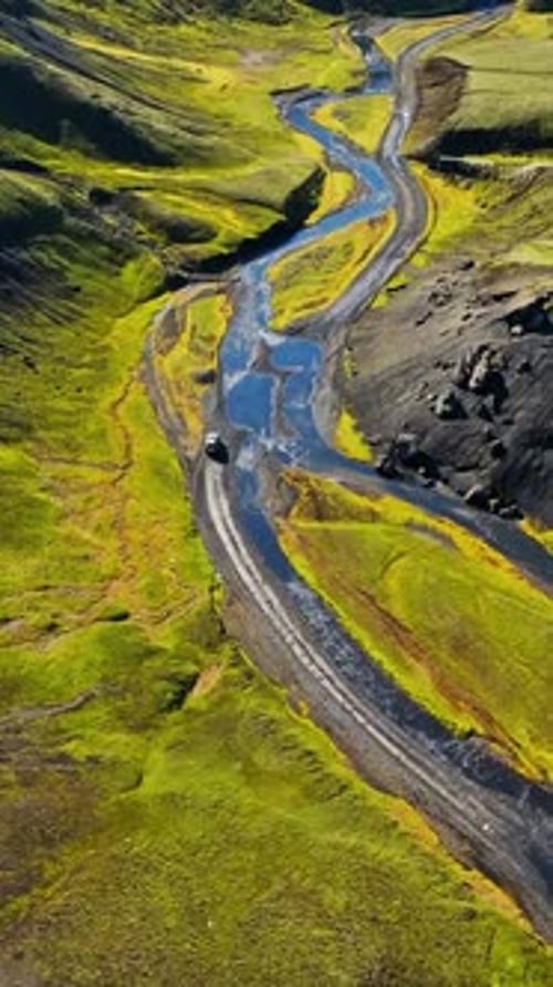 Vertical drone shot of a winding river beside a mountain road in the Iceland highlands, with a lone