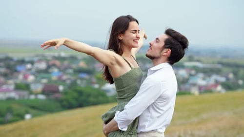 Young Man Holds Smiling Wife in Arms Kissing Against Village