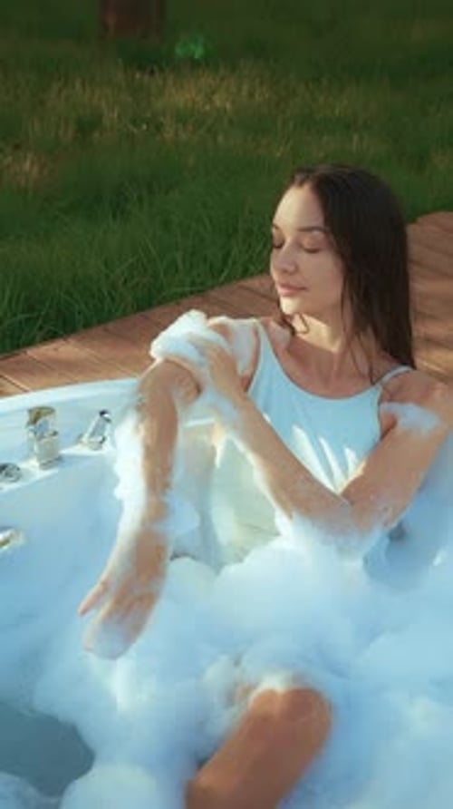 Woman enjoying a relaxing bubble bath outdoors surrounded by lush green grass and wooden decking