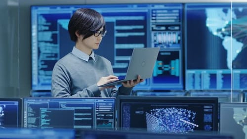 In the System Control Room IT Technician Holds and Works on a Laptop, in Background Multiple Displa