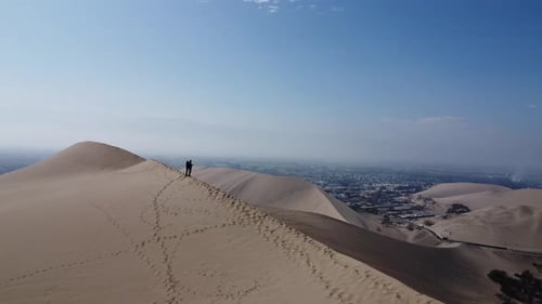 A pair of intrepid travelers stand on a dune near an oasis. Huacachina, Peru