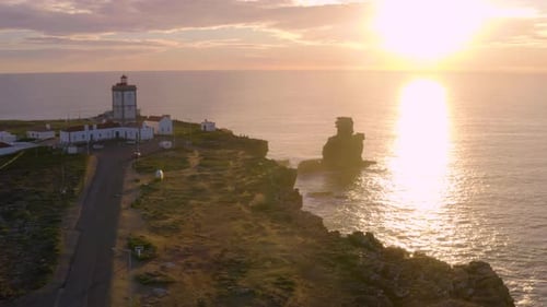 Aerial fly over coastal lighthouse with waves crashing on the cliffside.