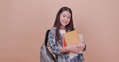 Cute school girl is carrying a school bag and holding a textbook on a brown background.