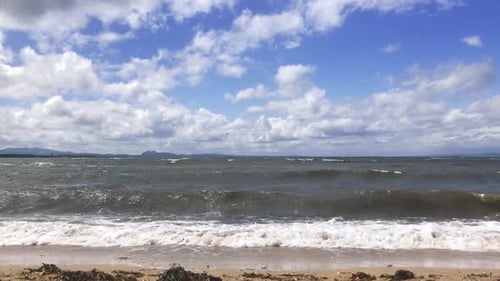 A fixed shot of waves breaking on a sandy beach with seaweed on the sand, on a windy summer day | Po