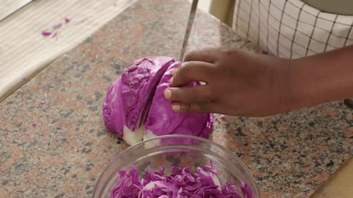 African man in apron placing halved purple cabbage on counter, shredding with chef knife for salad