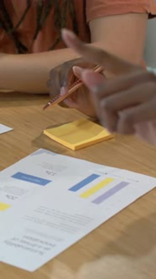 Vertical video: Leaning, duo pointing, tracing graph for review on desk with rose pen, coral top
