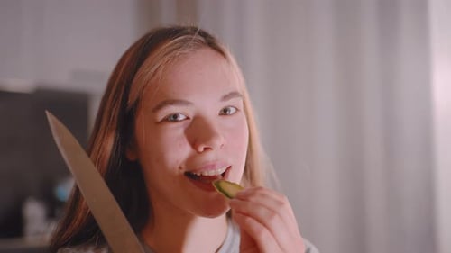 Young Woman Eating Cucumber Slices Holding Knife Indoors