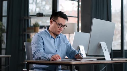 Serious Middle Aged Asian Man in Eyeglasses Working at Office Comparing Information on Computer