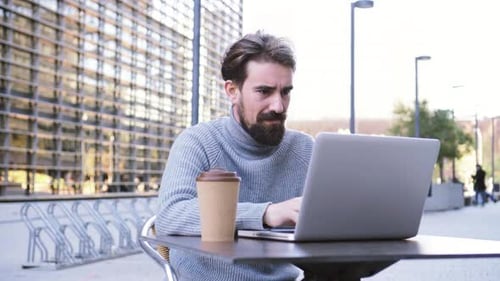 Unternehmer, der online mit einem Laptop-Computer arbeitet und draußen auf einer Stadtterrasse einen Kaffee trinkt