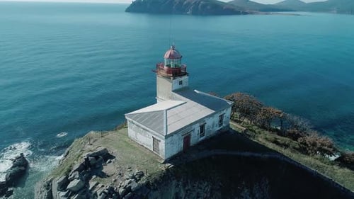 Aerial Panoramic View of White Lighthouse on the Rocky Cliff at Seaside Waves are Crashing to the