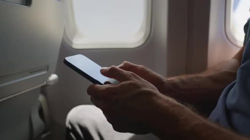 Man using smartphone mobile during flight on commercial airplane sitting next to plane window