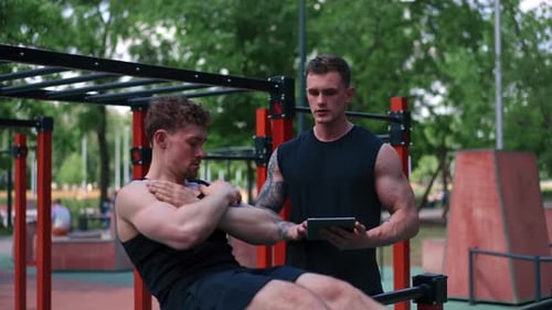 un entraîneur physique avec une tablette à la main entraîne un jeune athlète à pomper ses abdos dans la rue