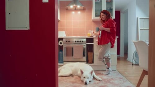 Happy Beautiful Woman with Cyber Bionic Leg Drinks Coffee Using Smart Phone in Light Cozy Kitchen