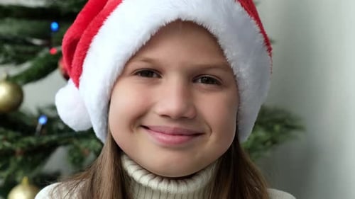 Close Up Smiling Kid Girl in Santa Hat on Background of Christmas Tree