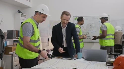 Construction Worker and Engineer Reviewing Blueprints in Office with Colleagues