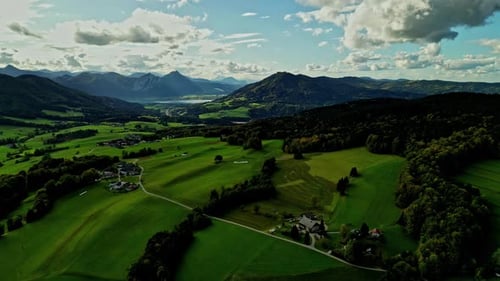 Lush green valley with scattered houses, flanked by mountains under a partly cloudy sky, aerial view