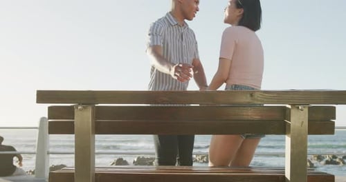 Happy biracial couple sitting on bench on promenade, in slow motion