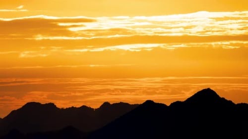 Silhouettes of mountains in golden sunset. Moving clouds in orange sky.