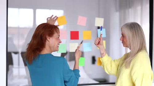 Professional businesswomen strategizing and brainstorming, using colorful sticky notes on glass wall