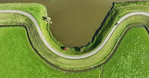 Aerial top down view of a car driving on a winding dam road through green fields and meadows.