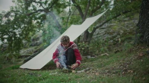 Man Making Campfire with Firewood beside Tarp Shelter in Forest