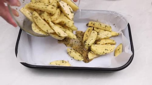 man pouring raw herb seasoned potato wedges for baking in a tray with parchment paper over white