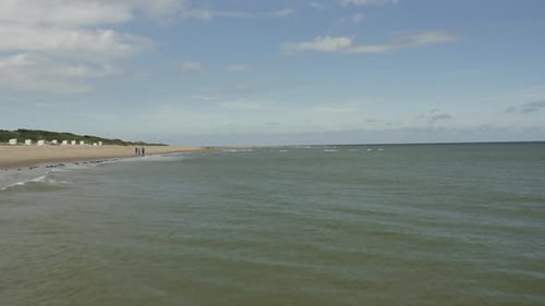 Aerial shot moving over waves at sea at high speed, along a beach where people are walking on a beau