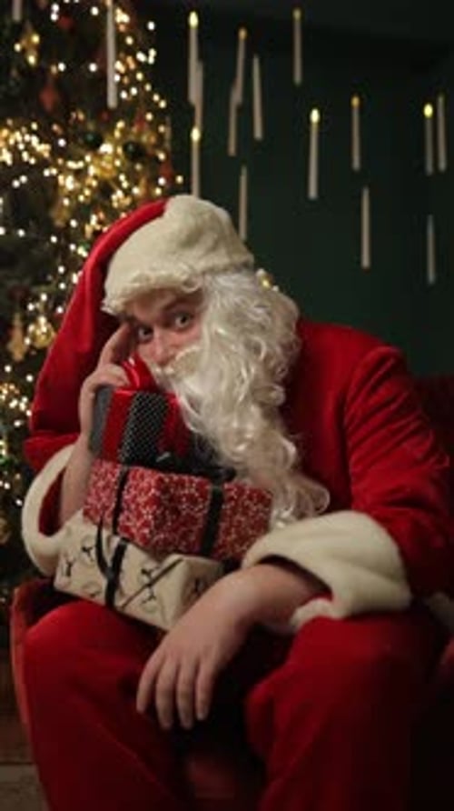 Vídeo vertical de Papai Noel cansado descansa abraçando uma grande caixa de presente de Natal perto da árvore