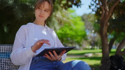 Young Woman Sitting on Park Bench Using Tablet While Looking Around with Thoughtful Expression Amid