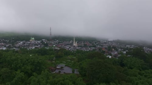 Aerial view of misty village, mosque, fields with mountain backdrop, Indonesia.