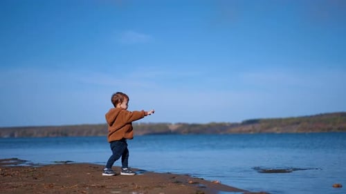 Little toddler boy approaching the river. Caucasian kid throws two stones into water.