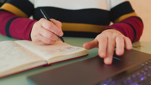 Person Writing in Notebook with Laptop Computer