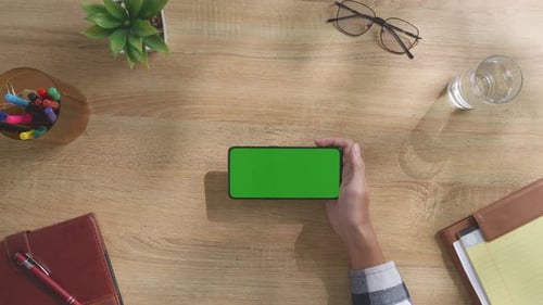 Top down view of man's hand holding mock up green screen smartphone on a desk
