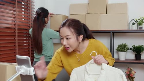 Two young asian woman live online clothing store while showing clothing with tablet on social.