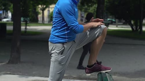 Young, Sporty Man Stretching Legs on Bench in City Park 30s