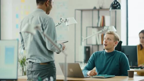 Hübscher IT-Spezialist, der an seinem Schreibtisch sitzt, arbeitet an einem Laptop und spricht mit einem professionellen Projektmanager