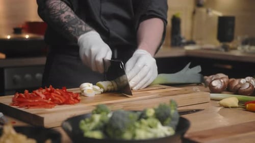 Fresh green leek being cut on a wooden board by young professional male chef in an elegant black shi