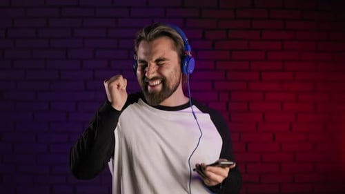 Positive Handsome Guy Using Modern Smartphone and Headphones for Listening Favorite Music in Studio