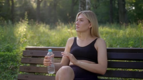 Nice Young Woman Sits on Bench in Park Forest in Morning After Jogging and Drinks Water