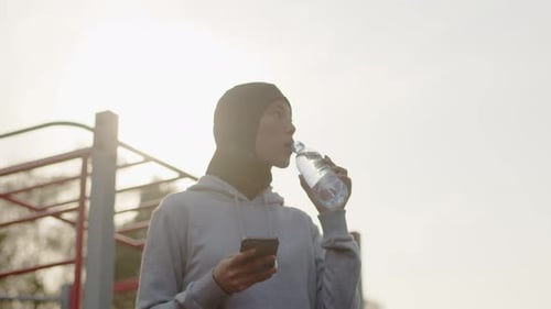Young Asian Muslim Woman in Hijab Looking at Her Smartphone and Drinking Water From the Bottle Close