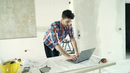 Man Using Laptop with Blueprints on Table