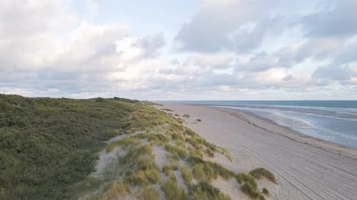 Slow aerial flyover sandy grass dunes beside beach and blue water of north sea in Netherlands