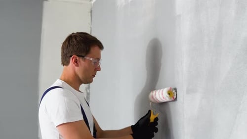 Side View of Happy Young Male Painter in Blue Overall and Gloves Painting Wall with Roller in Grey