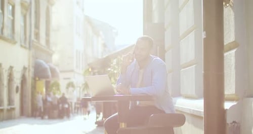 Handsome Young Man Sitting at Table of Cafe in City Center with Laptop and Talking on Mobile Phone