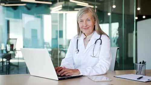 Portrait of smiling senior gray haired female doctor in white coat working on laptop in hospital